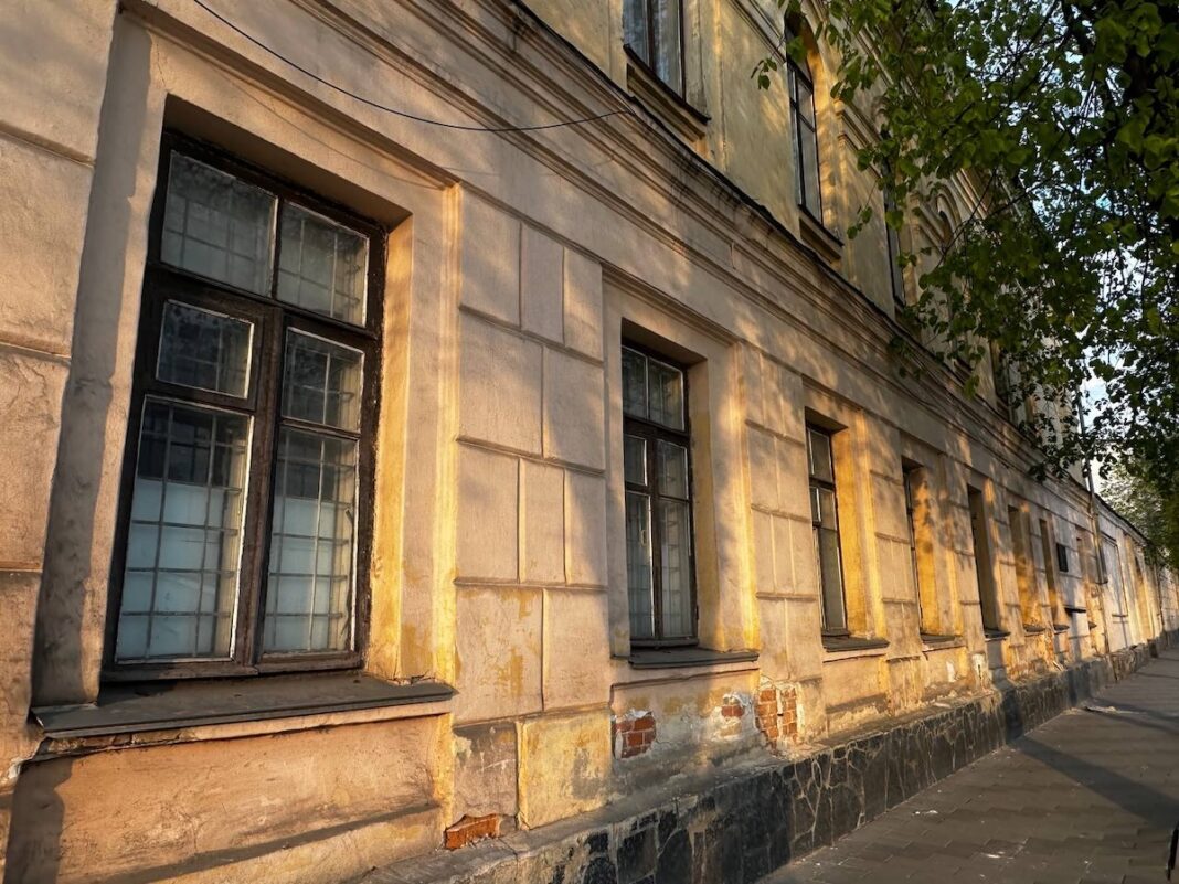 Пошкоджений фасад двоповерхового старовинного будинку жовто-бежевого кольору з сильно облупленою штукатуркою на цоколі та оголеною червоною цеглою