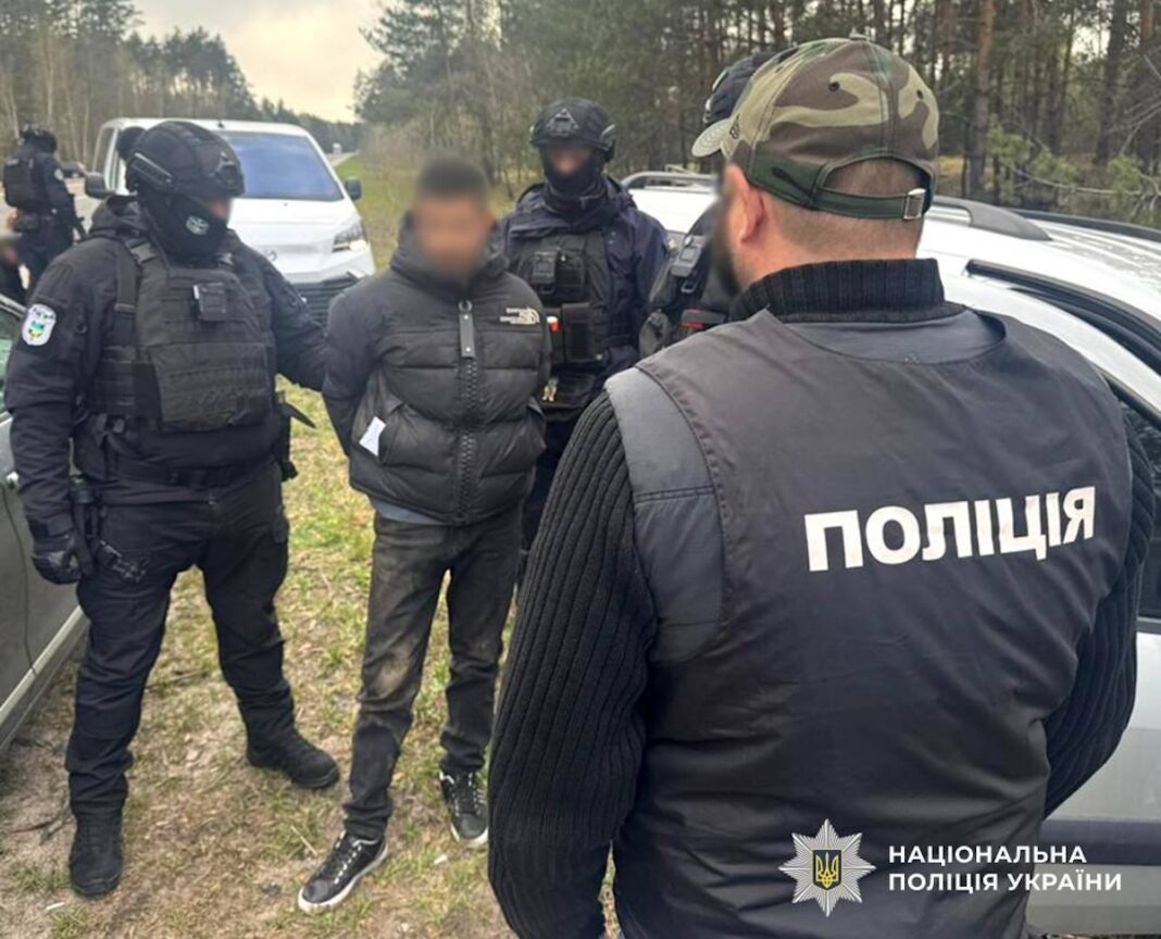 Затриманий чоловік у чорному одязі зі схованими за спиною руками стоїть поруч із двома озброєними бійцями спецпідрозділу поліції у балаклавах на узбіччі