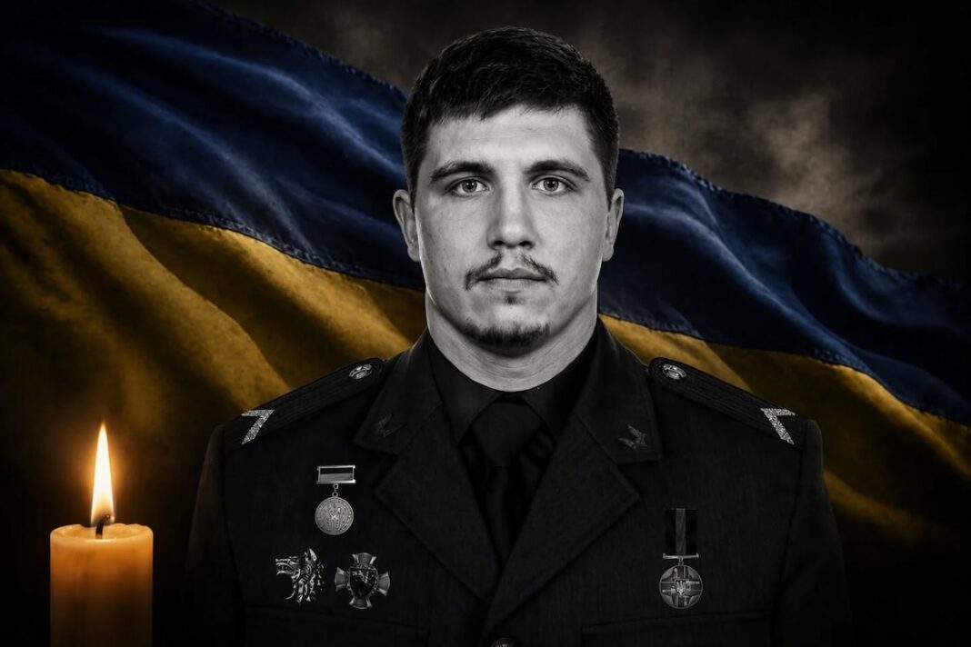 Портрет молодого загиблого українського військовослужбовця у чорному кітелі з нагородами на тлі синьо-жовтого прапора та запаленої свічки