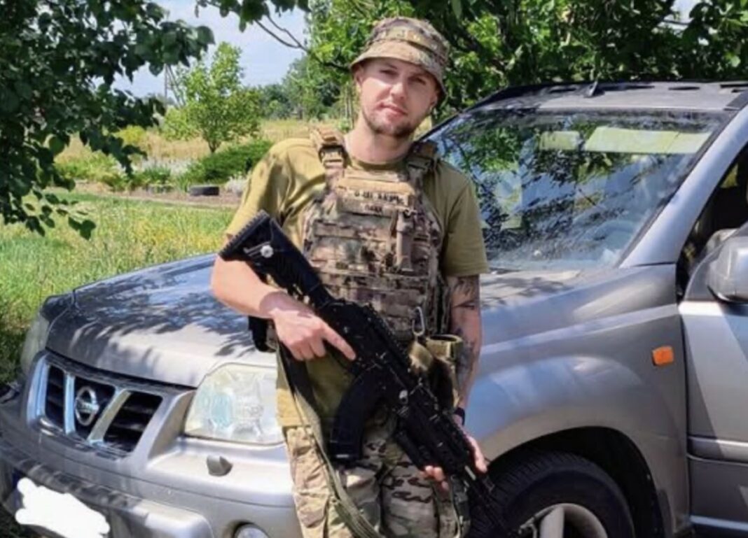 Захисник України Павло Білоус у військовій екіпіровці зі зброєю в руках стоїть біля позашляховика