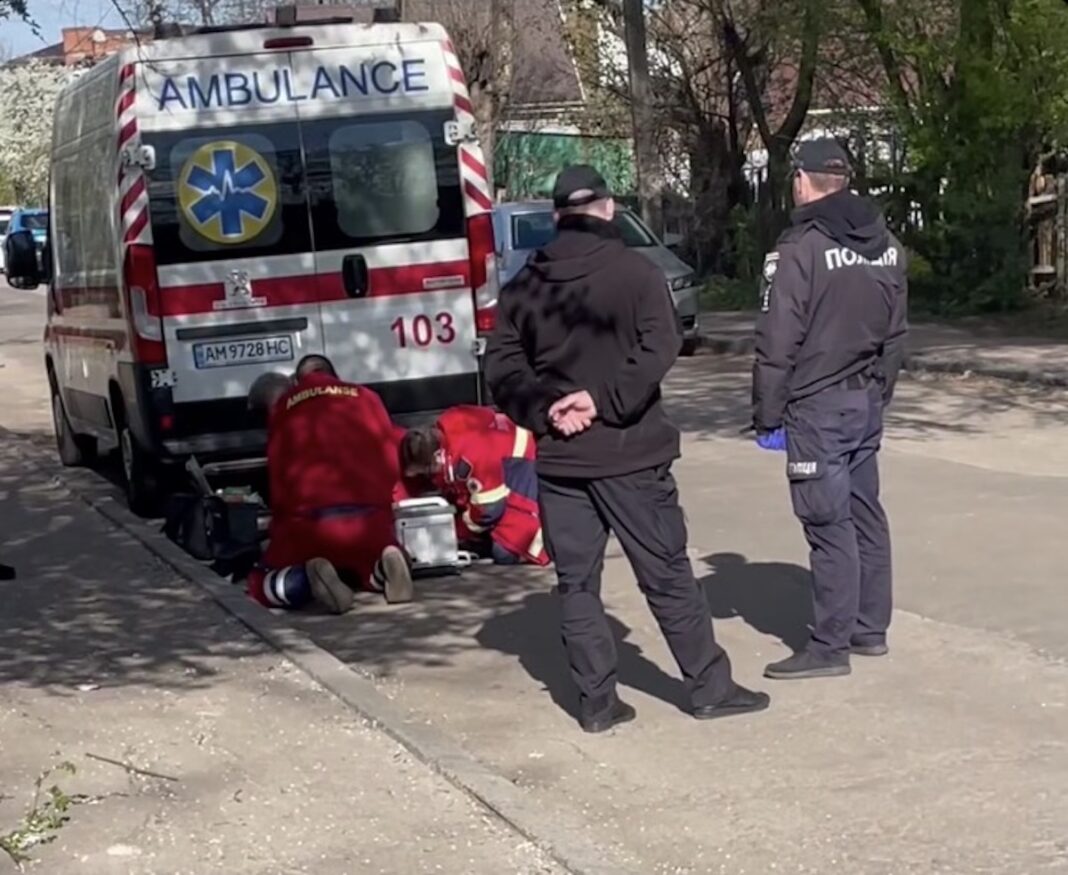 Двоє медиків у червоній формі проводять реанімацію людині на асфальті біля карети швидкої, за ними спостерігають поліцейські