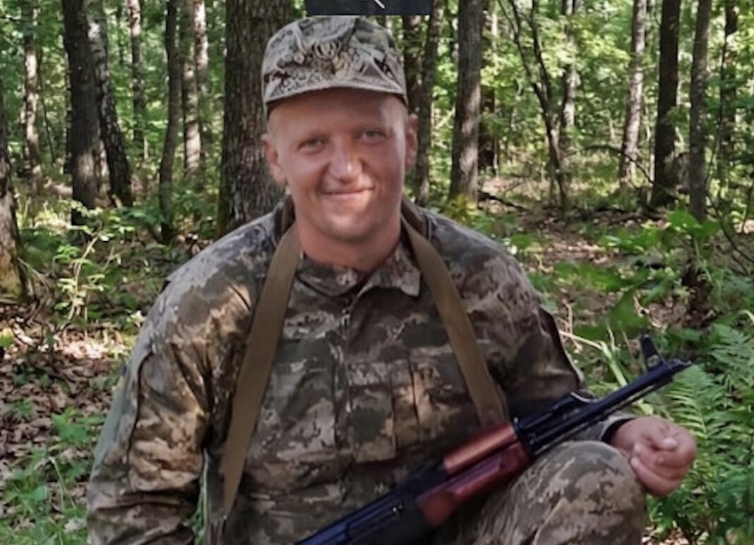 Український військовослужбовець Вячеслав Лісовий у камуфляжній формі та польовій кепці привітно посміхається, тримаючи в руках автомат на тлі лісу