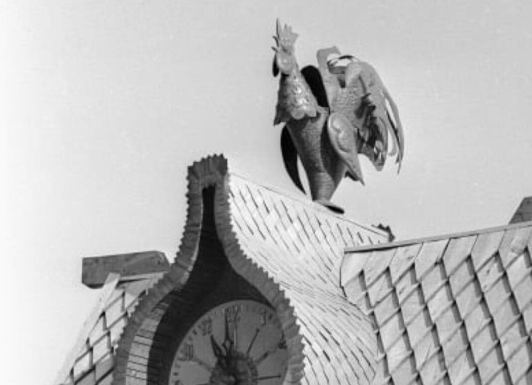 Архівне чорно-біле фото фігурного даху з великим циферблатом годинника, над яким встановлена деталізована мідна фігура півня