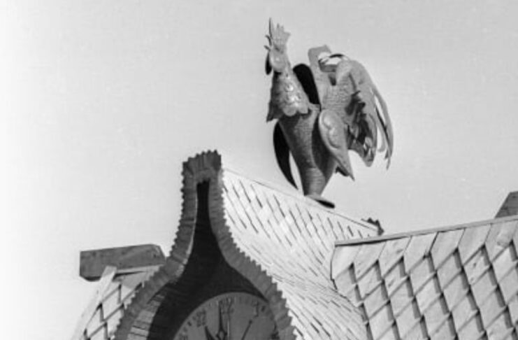Архівне чорно-біле фото фігурного даху з великим циферблатом годинника, над яким встановлена деталізована мідна фігура півня