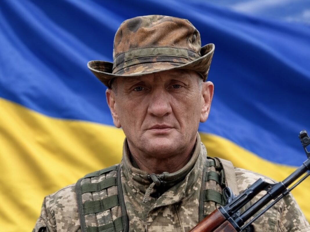 Український військовослужбовець Василь Прокопенко у камуфляжній формі та тактичній панамі тримає в руках зброю на тлі державного прапора