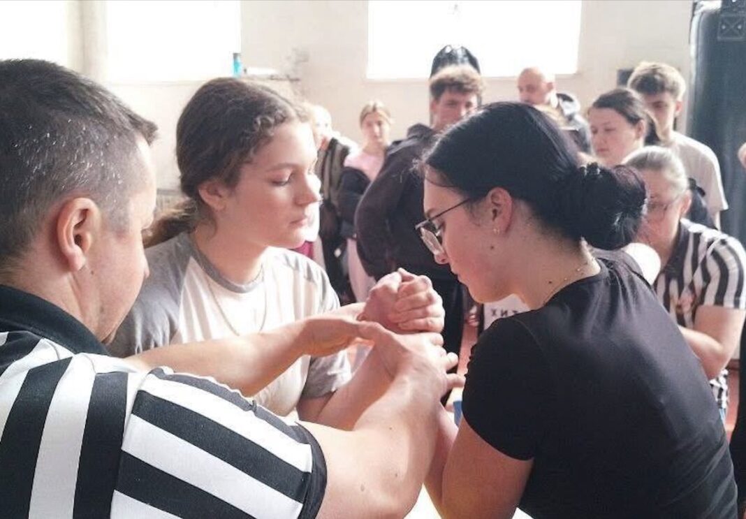 Дві дівчини змагаються за столом для армрестлінгу, суддя у смугастій чорно-білій формі контролює їхній стартовий захват