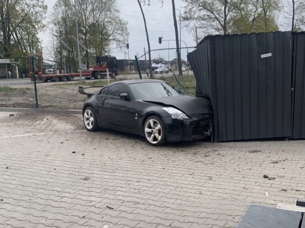 Чорний спортивний автомобіль Nissan 350Z із заднім спойлером стоїть на тротуарній плитці, врізавшись передньою частиною у конструкцію з металевого профнастилу