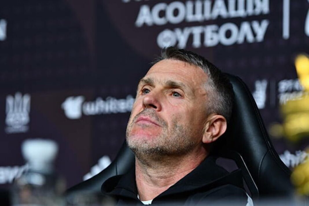 Сергій Ребров із зосередженим виразом обличчя сидить у чорному кріслі під час пресконференції на тлі офіційного банера УАФ