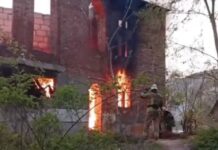 Рятувальник у захисному шоломі та формі стоїть спиною до камери перед цегляною двоповерховою недобудовою, всередині якої вирує яскраве полум'я